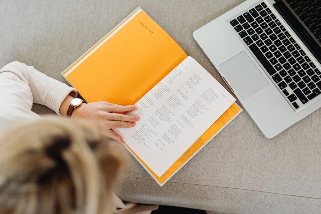 Woman reading a book with laptop nearby, showcasing technology and leisure blend.