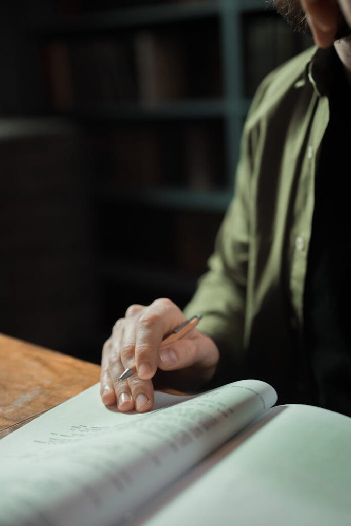A man writes on a notebook at a wooden table in a dimly lit library.
