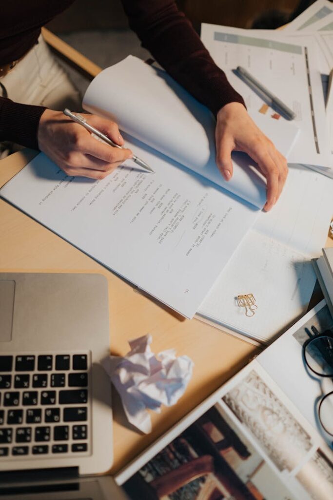 Woman writing in a notebook surrounded by papers and a laptop, showcasing a productive work environment.